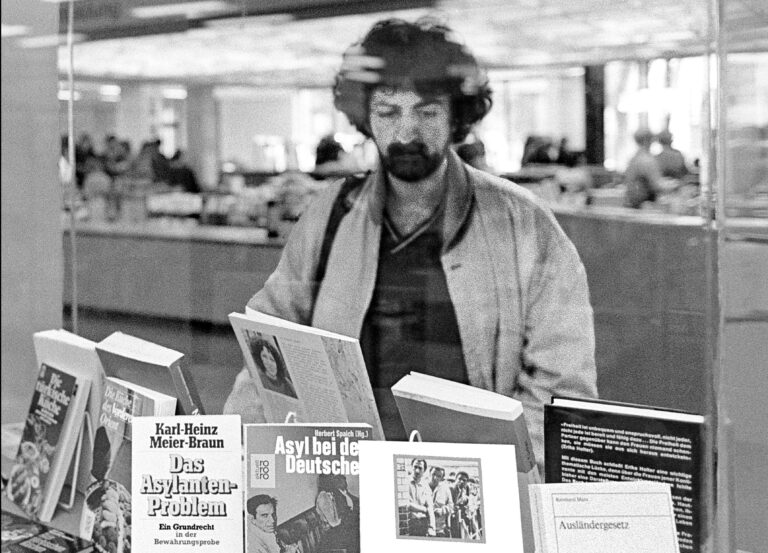 80&#39;li yıllardan siyah beyaz bir fotoğraf. Üzerinde &quot;Sığınmacı Sorunu&quot; gibi başlıklar bulunan bir kitap masasının önünde Türk asıllı bir adam duruyor. 