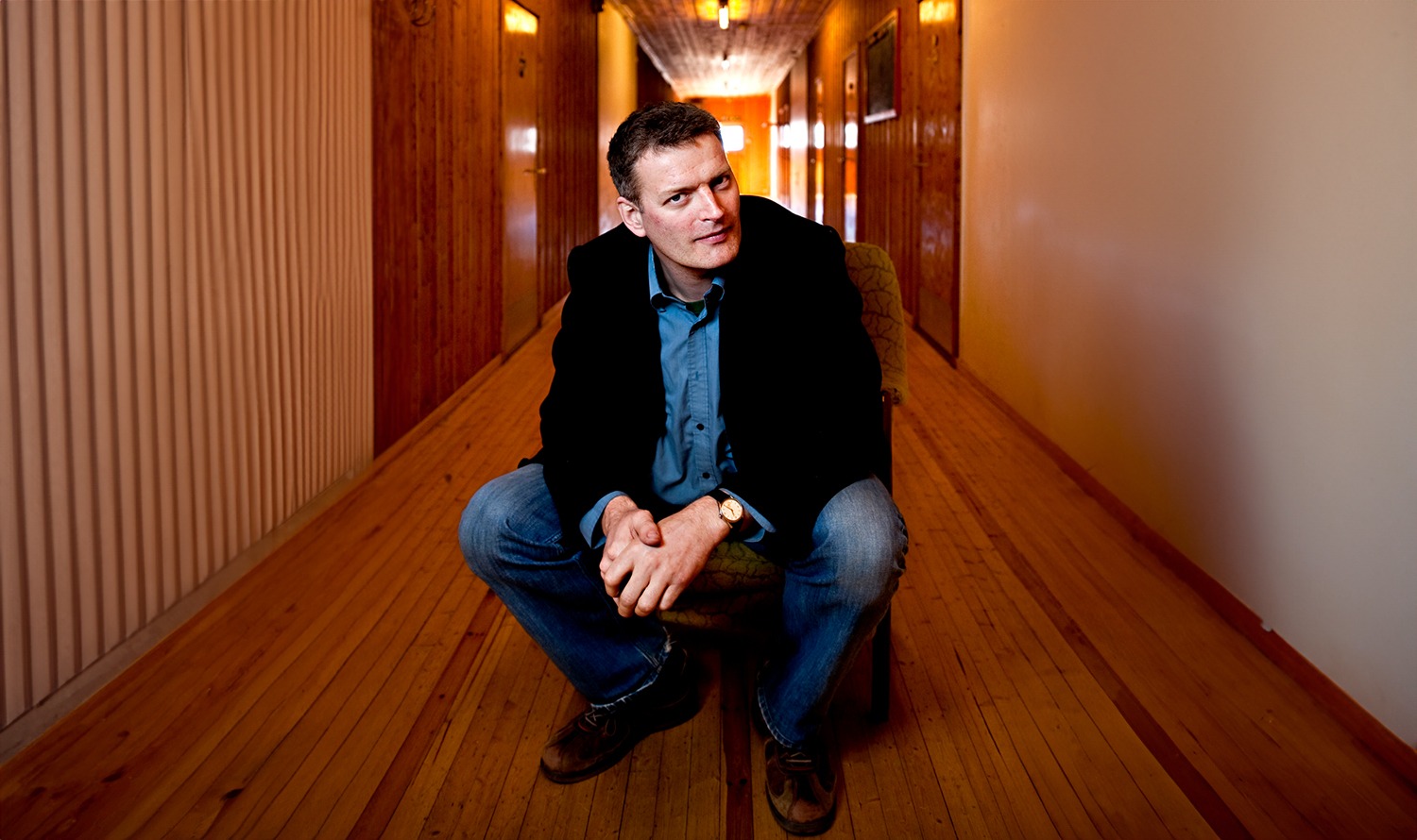 Portrait: Lars Mytting in a long hallway with old wooden floorboards, doors and a wooden ceiling, surrounded by warm light. It conveys an atmosphere of comfort and tradition. 