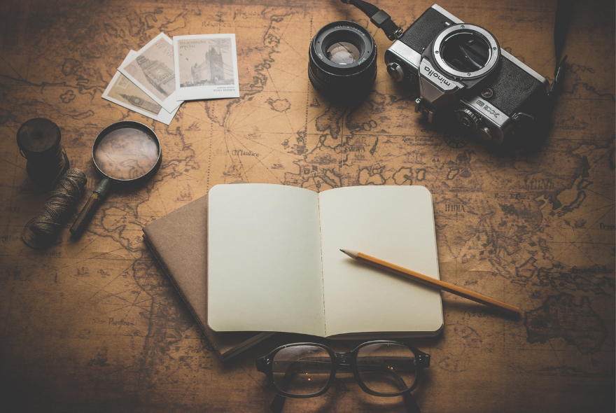 Camera, magnifying glass, glasses, pen and empty notebook lie on a map.