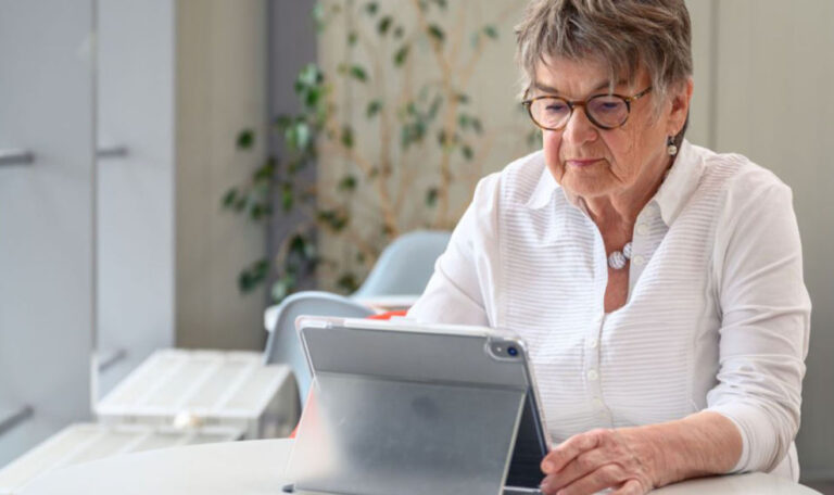 Пожилая женщина сидит за столом и работает с планшетом.