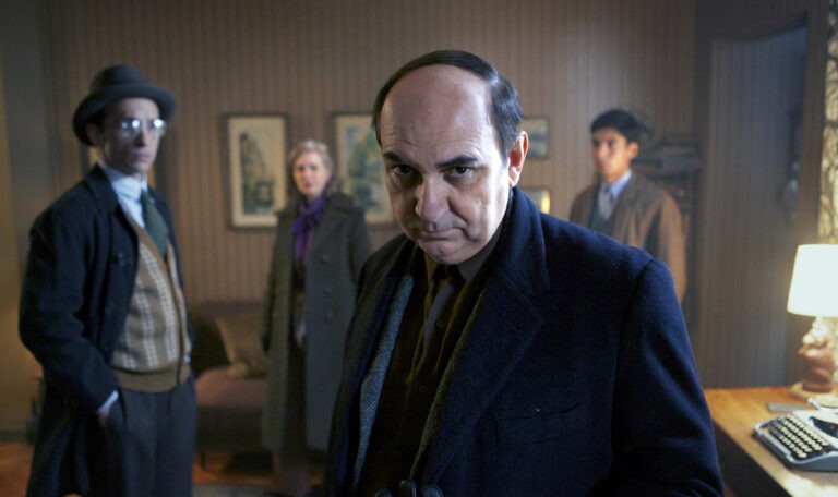 The image shows four people in a room, with the man in the foreground looking into the camera with a serious expression, while the other three people in the background remain out of focus.