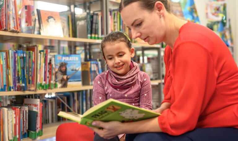 O femeie îi citește o carte unei fete într-o bibliotecă, în timp ce amândoi zâmbesc.