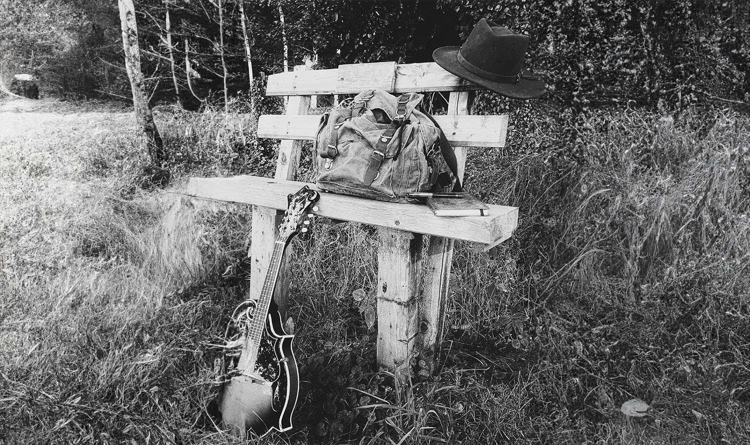 The image shows a rustic outdoor wooden bench with a hat, backpack, notebook and mandolin placed on it.