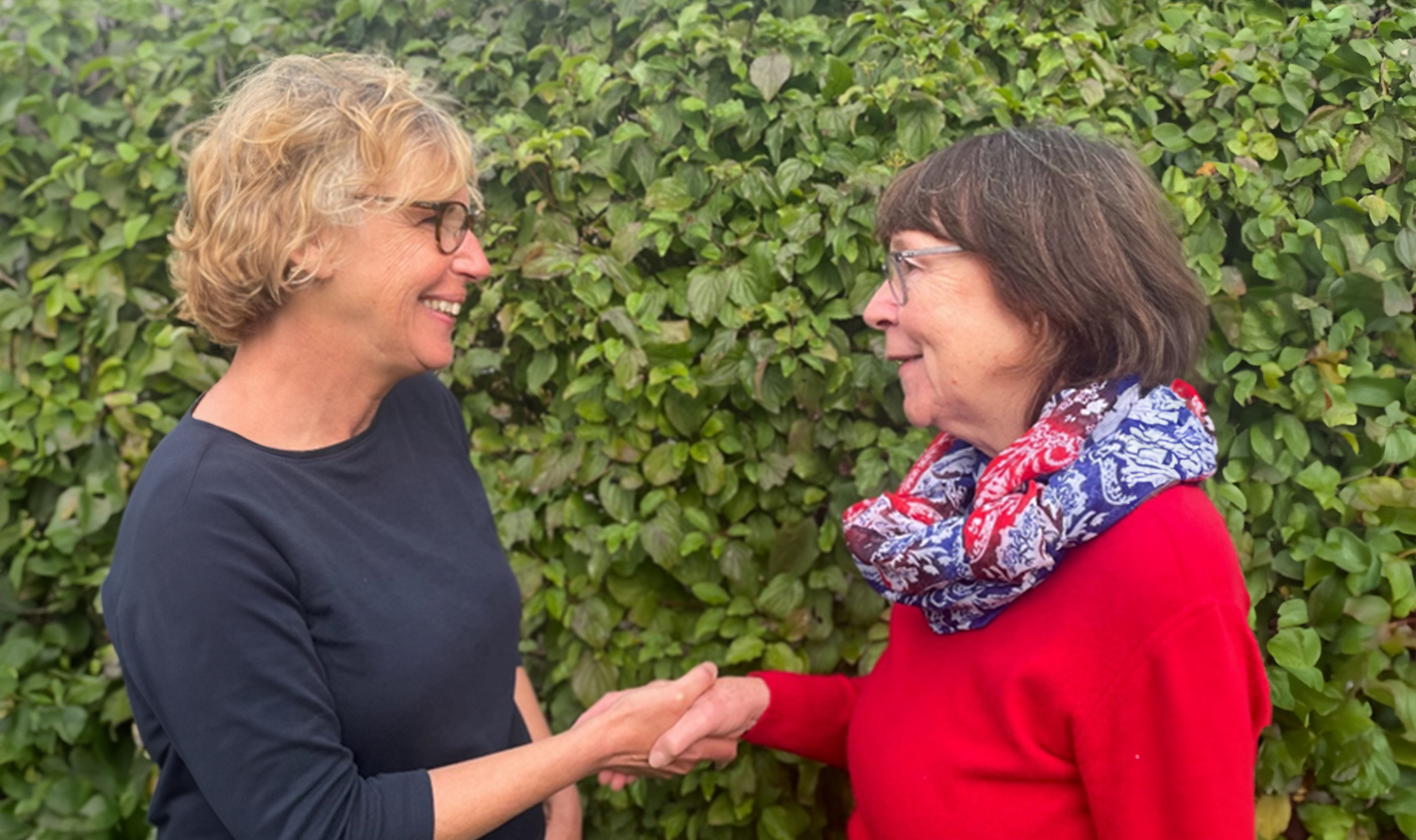 The picture shows two women shaking hands and smiling at each other, with a green hedge in the background.